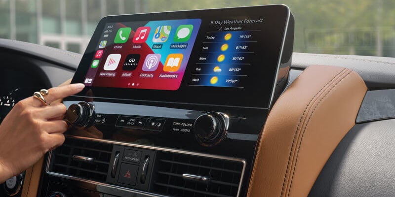 Hand touching a large infotainment screen displaying Apple CarPlay and a weather forecast.