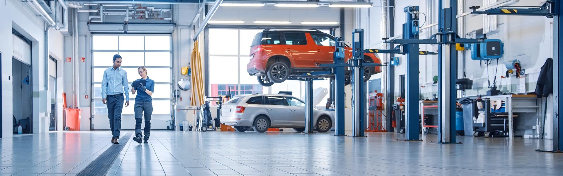 Two people walk through a brightly lit car service center with a lifted orange car and other tools.