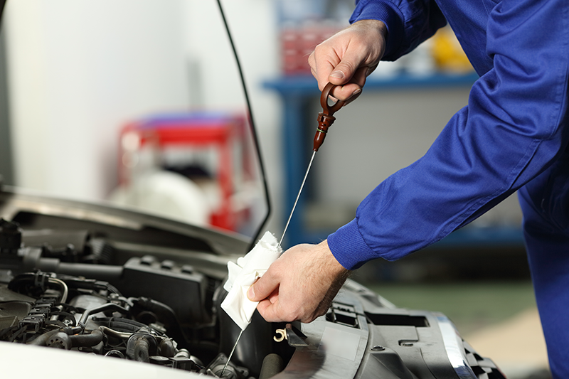 Mechanic in blue uniform checking engine oil level with a dipstick under the hood of a car in a workshop.